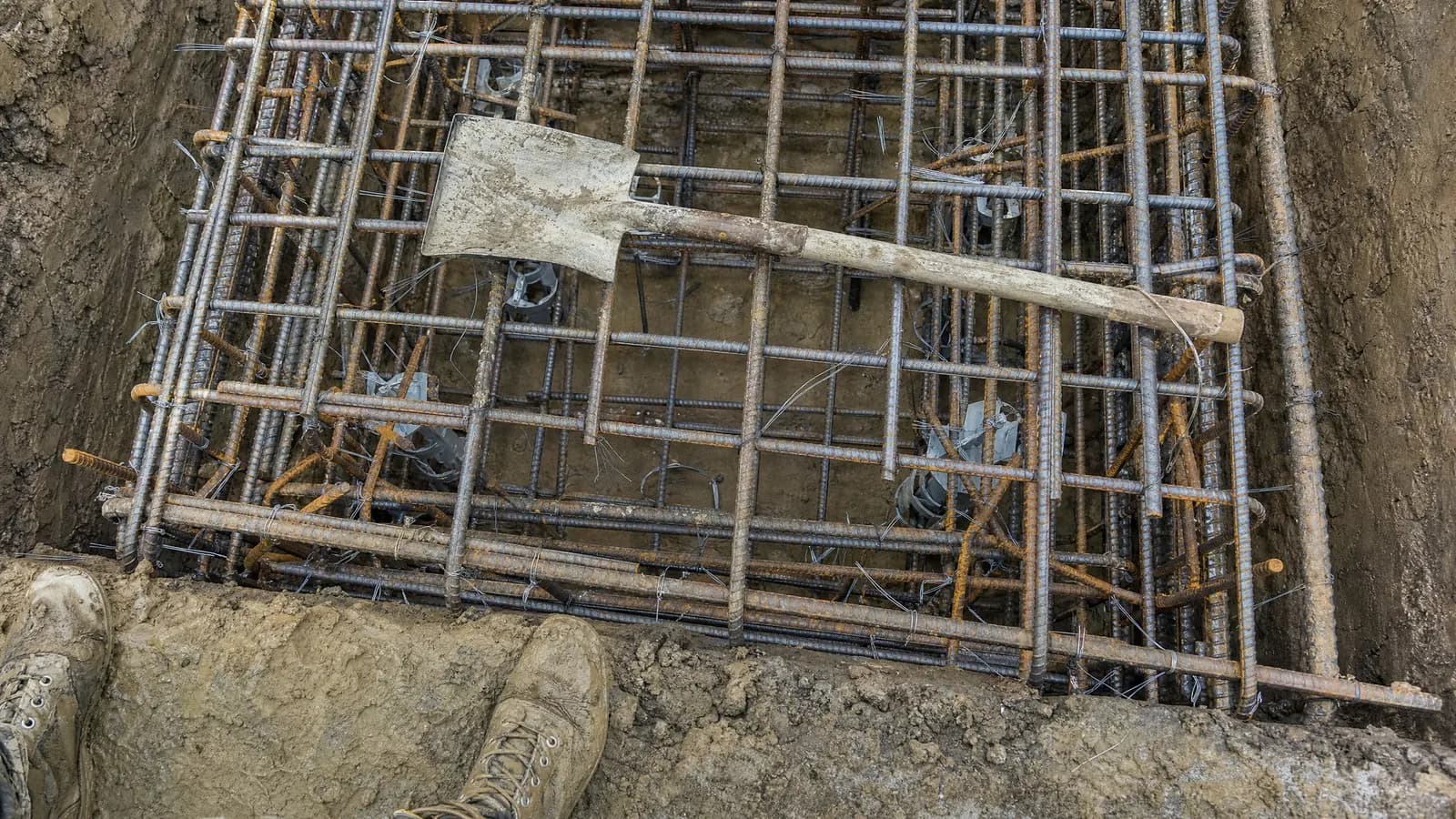 Looking down into a foundation trench at a dense cage of tied reinforcing steel ready for a concrete pour, with muddy boots at the edge of the frame.