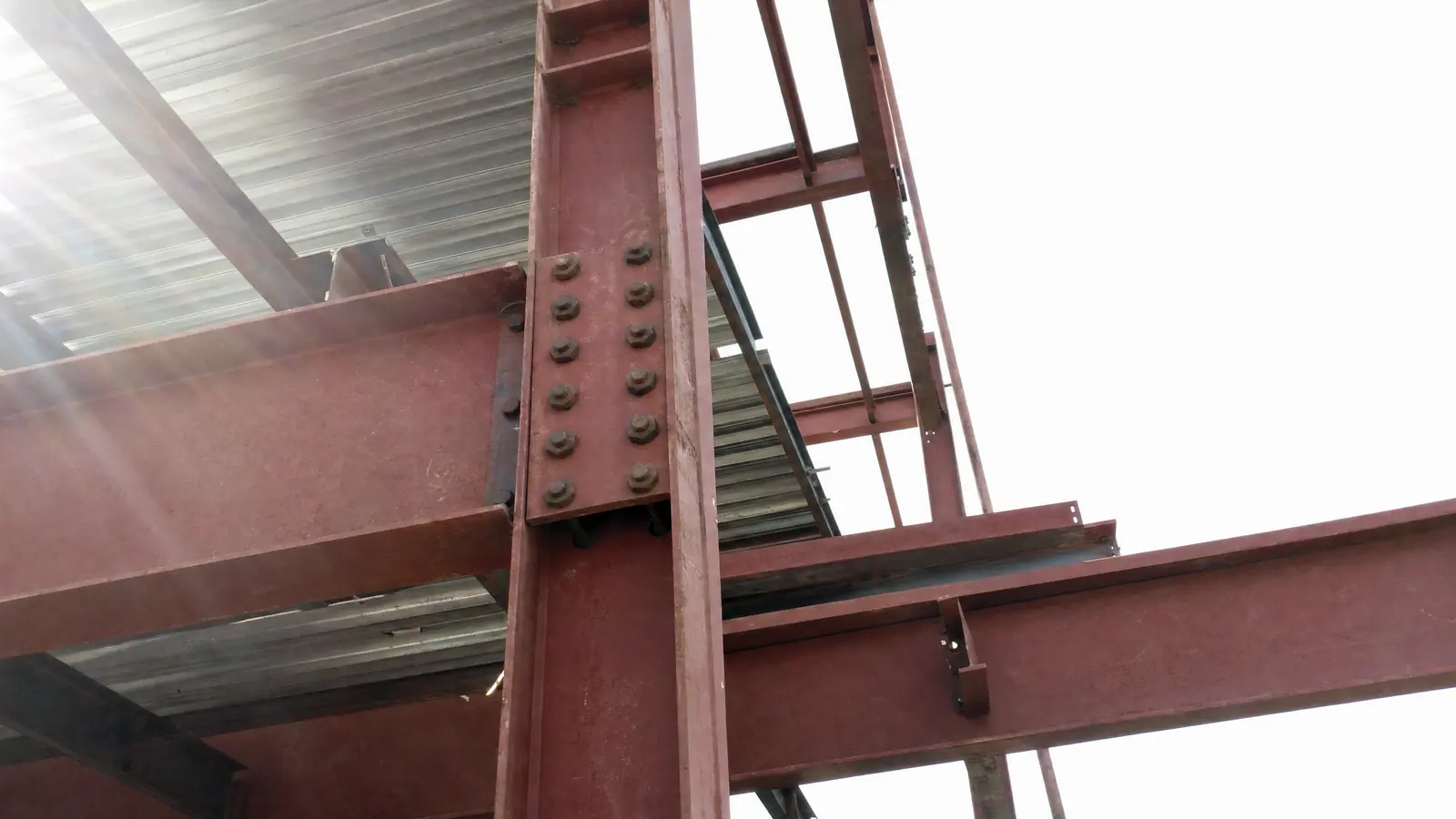 A structural steel frame photographed from below against a bright sky, a bolted beam connection close to the camera and metal decking part-laid above.