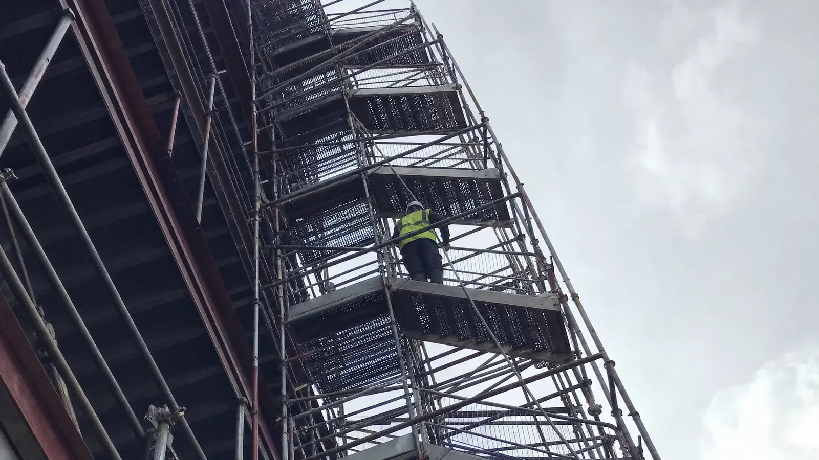 Looking steeply up an exterior scaffold stair tower as a lone worker in a hard hat climbs several flights above, the stairs receding into a bright sky.