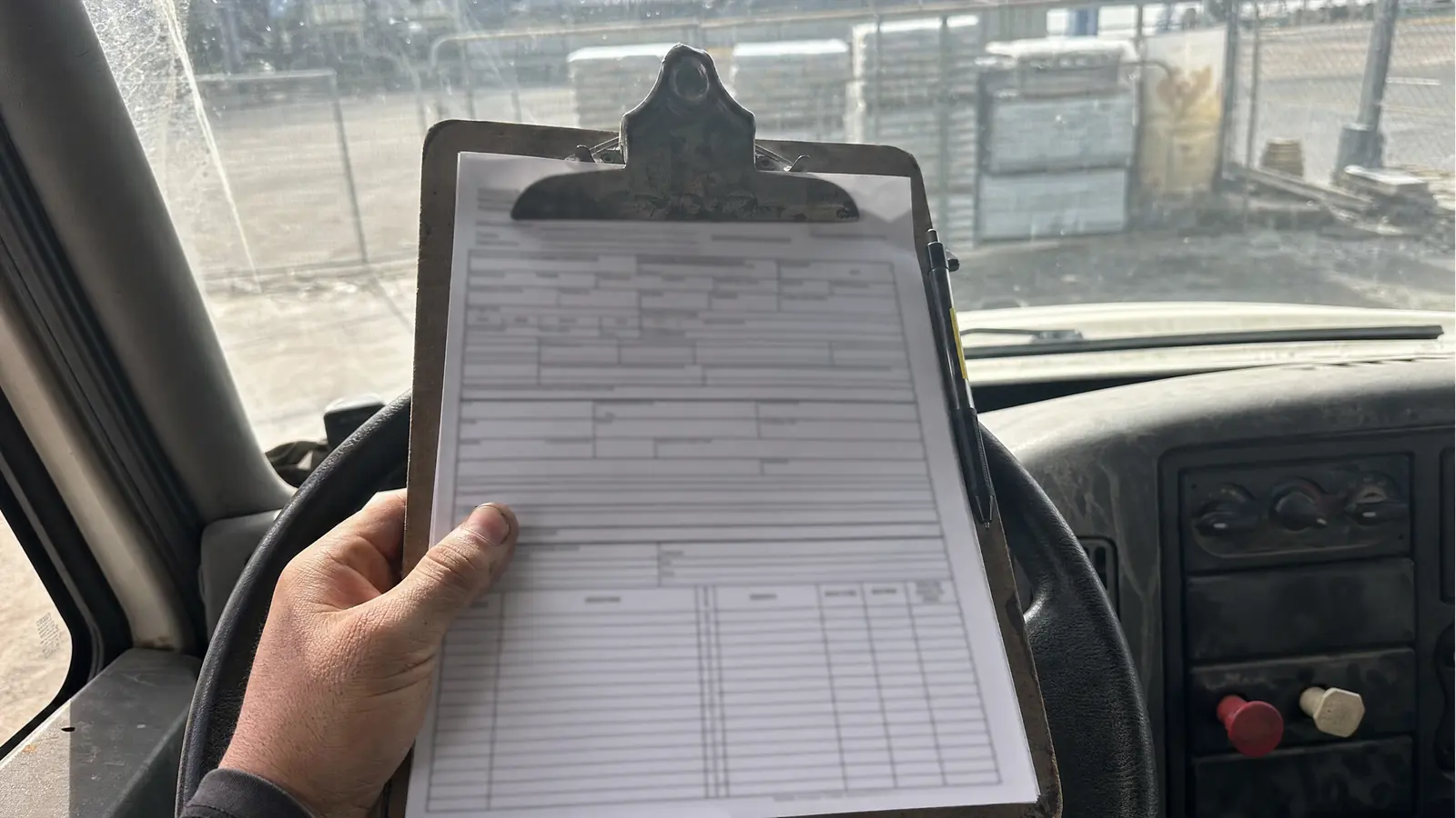 A clipboard of paperwork resting on a work truck's steering wheel, held down by a thumb, with a jobsite gate blurred through the windscreen beyond.