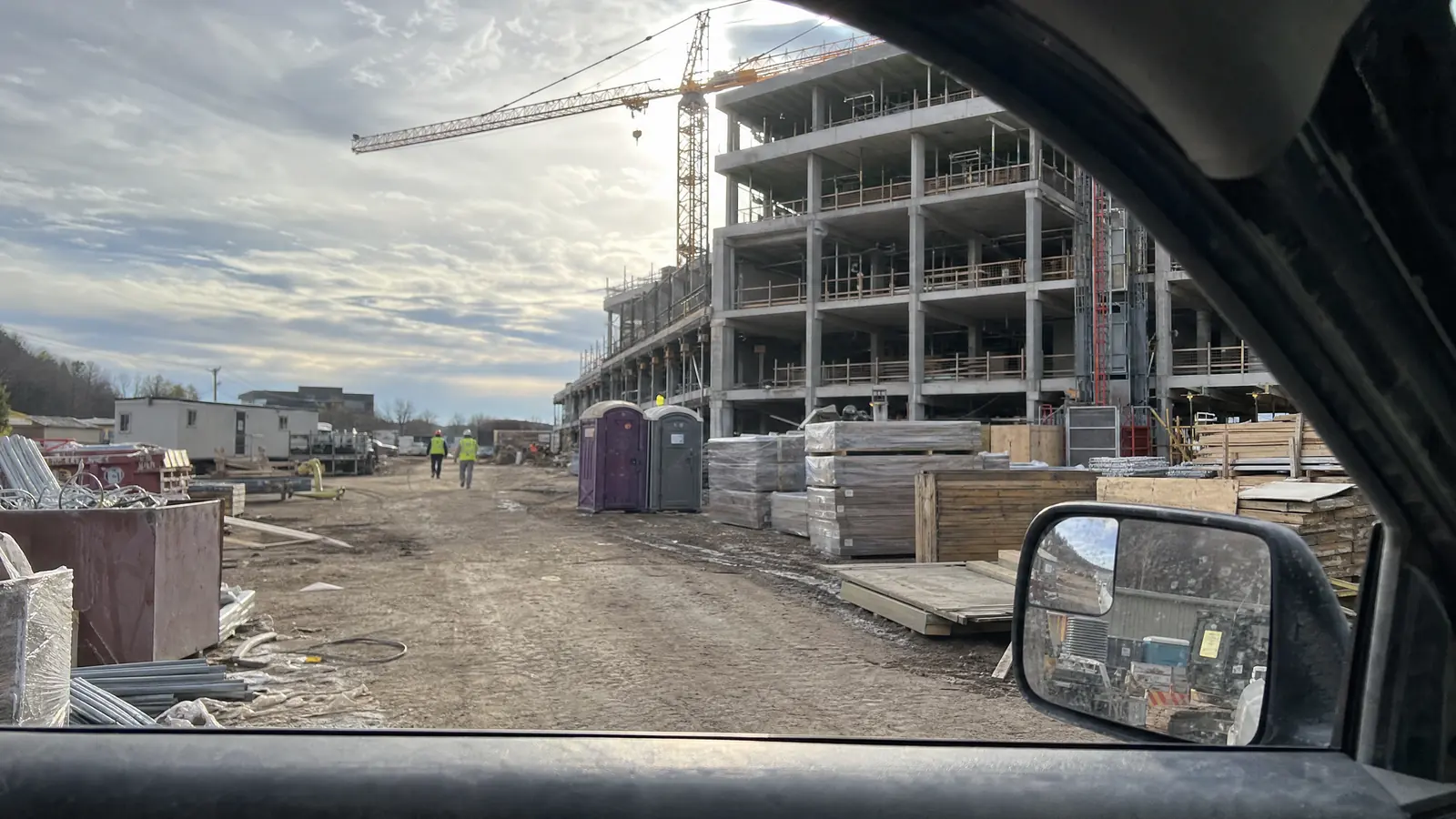An active commercial construction site in late afternoon, seen from a parked truck: a concrete frame several storeys up, a tower crane and site trailers.