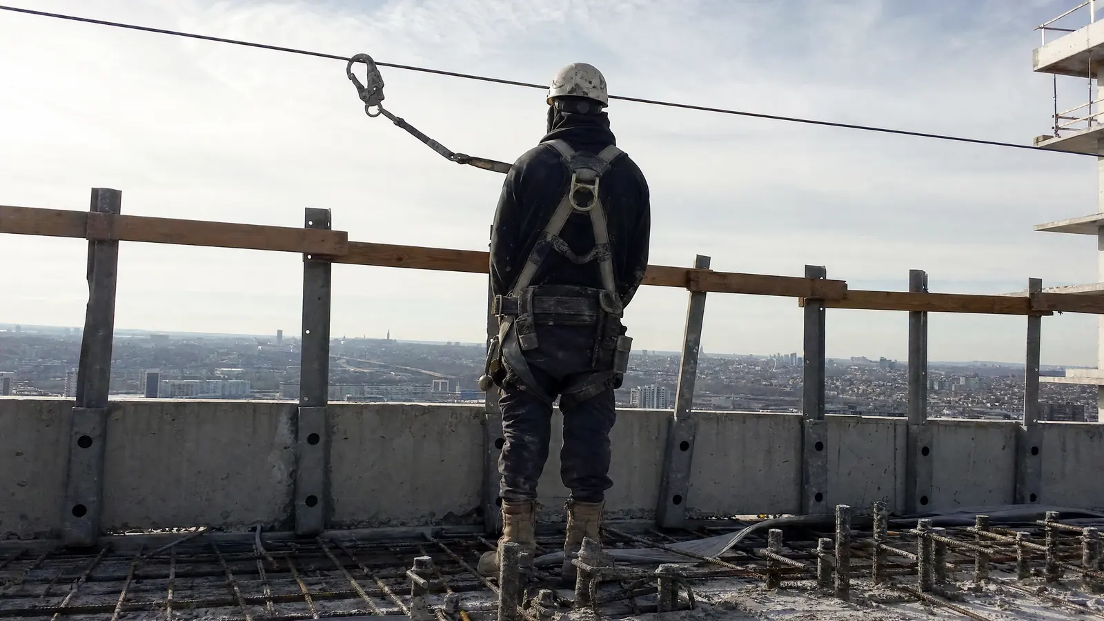 A worker seen from behind at the open edge of an upper concrete deck, their harness lanyard clipped to a taut horizontal lifeline.