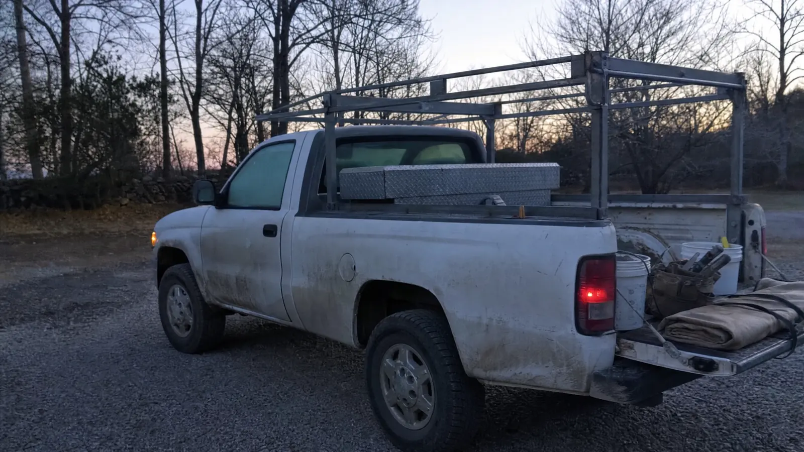 A work pickup with a ladder rack parked on a gravel drive at first light, tailgate down, the bed organised with a job box and buckets of tools.