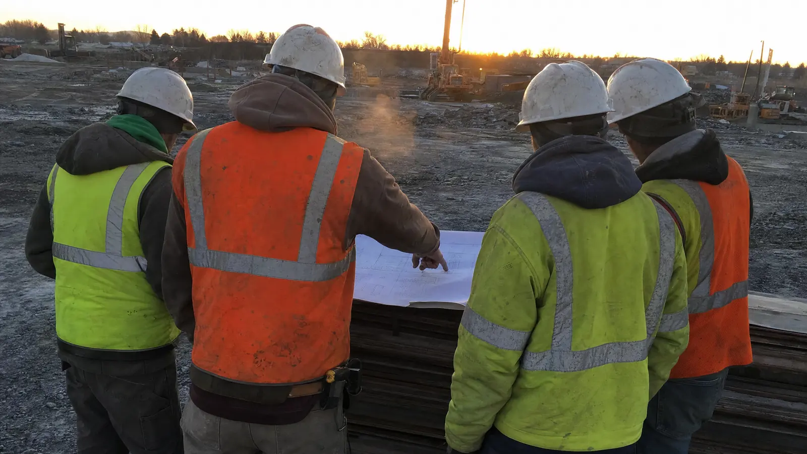 Seen from behind: four workers in vests and hard hats gathered around drawings spread on a stack of material, one pointing down at the sheet.