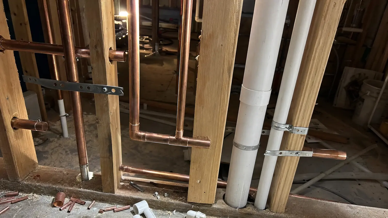 Plumbing rough-in inside an open stud wall: soldered copper supply lines, a white PVC waste stack and vent, and pipe strapping through drilled studs.