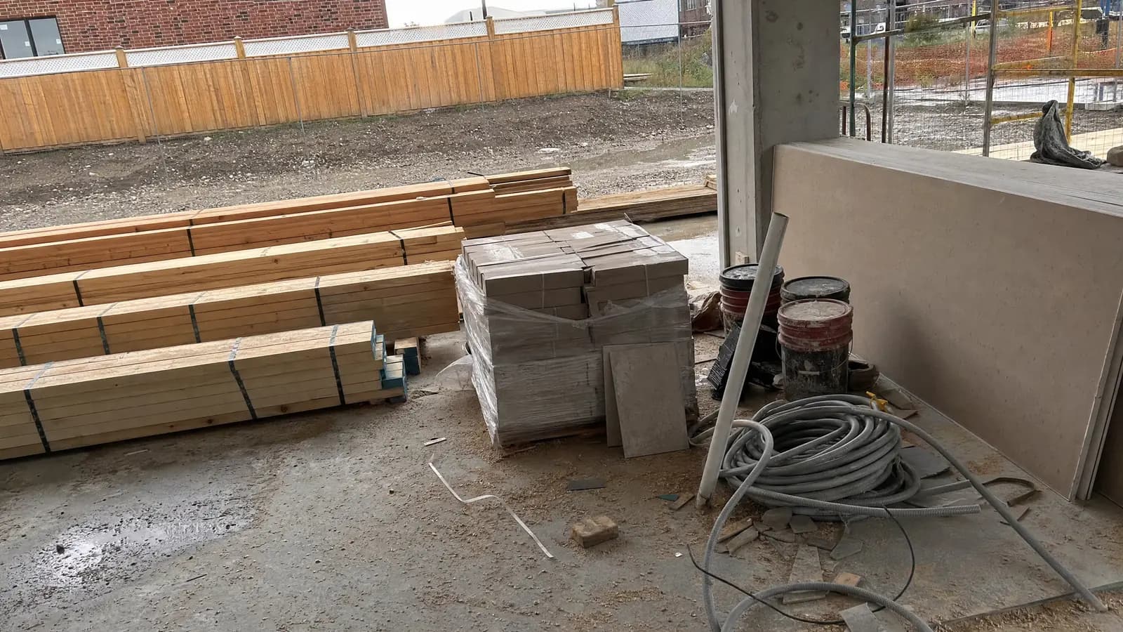 Building materials staged on a bare concrete slab: banded timber studs, a shrink-wrapped pallet of tile, coiled conduit and leaning drywall sheets.