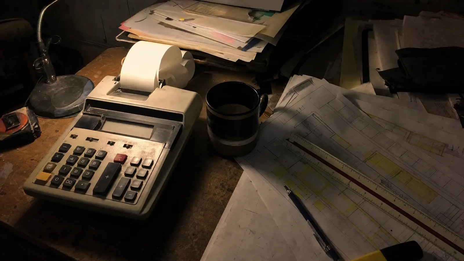 A desk at night under one clamp light: a calculator with a curling paper tape, a scale rule, and drawings marked up with yellow highlighter.