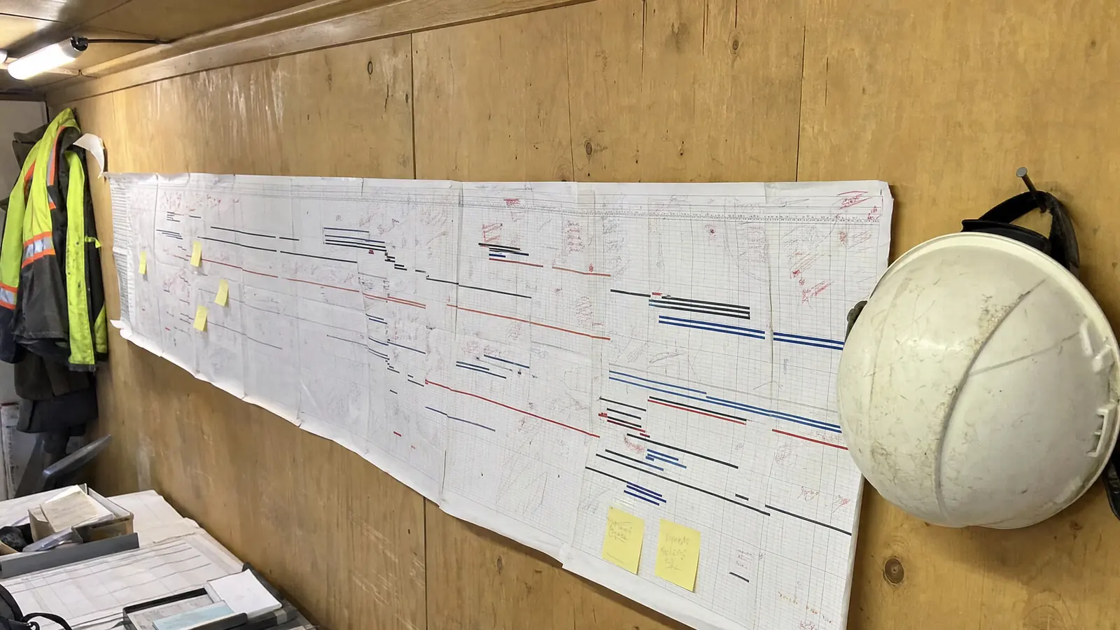 A long printed bar-chart schedule taped across a site-trailer wall, joined from several sheets and covered in handwritten red-pen marks.