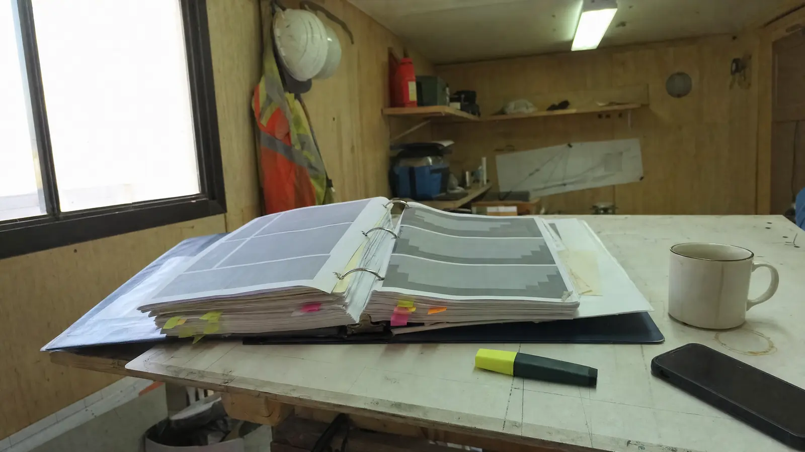 A thick specification binder open on a cluttered site-trailer table, marked with dozens of coloured sticky tabs, beside a highlighter and a coffee mug.