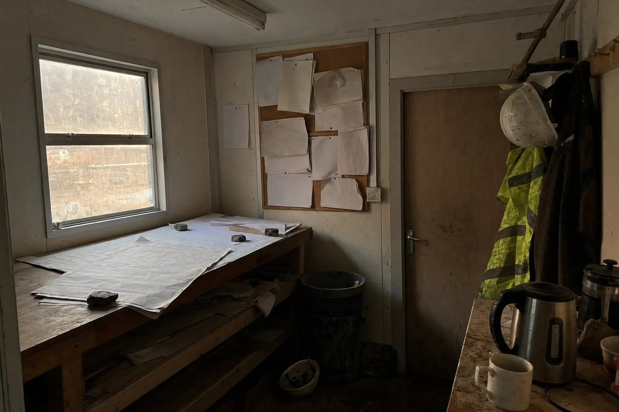 The inside of a site trailer at the end of the day: drawings weighted on a plan table, a corkboard of pinned paper, and a hard hat and vest on hooks.