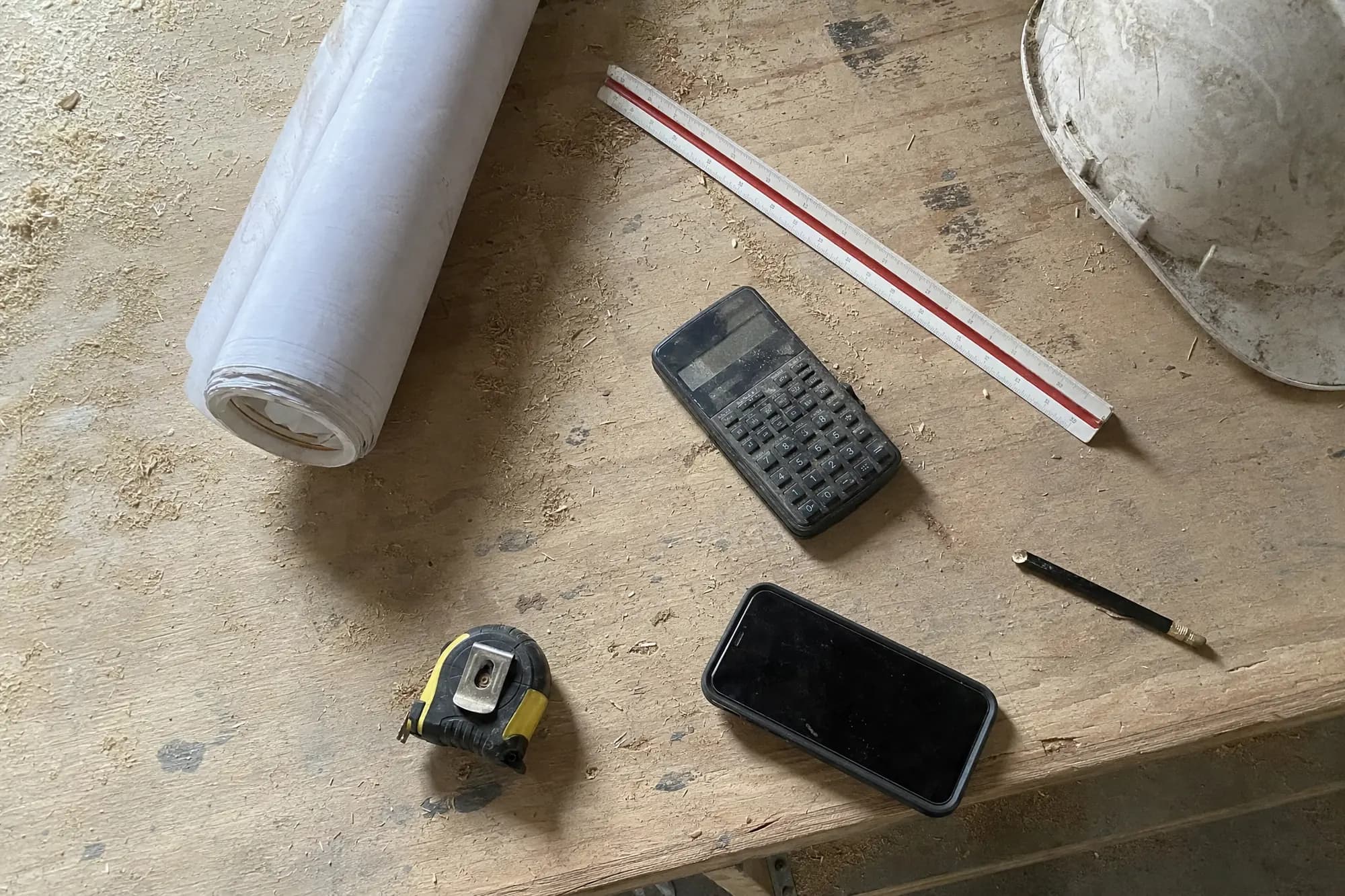 An overhead view of a scarred plywood workbench with rolled drawings, a scale rule, a calculator, a tape measure and a hard hat scattered across it.